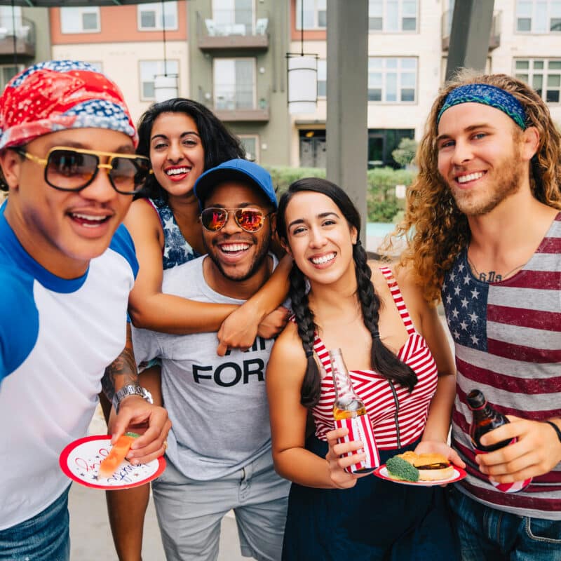 Group of five friends posing for a selfie at an outdoor pool, wearing red, white, and blue outfits and holding snacks/drinks in hand.