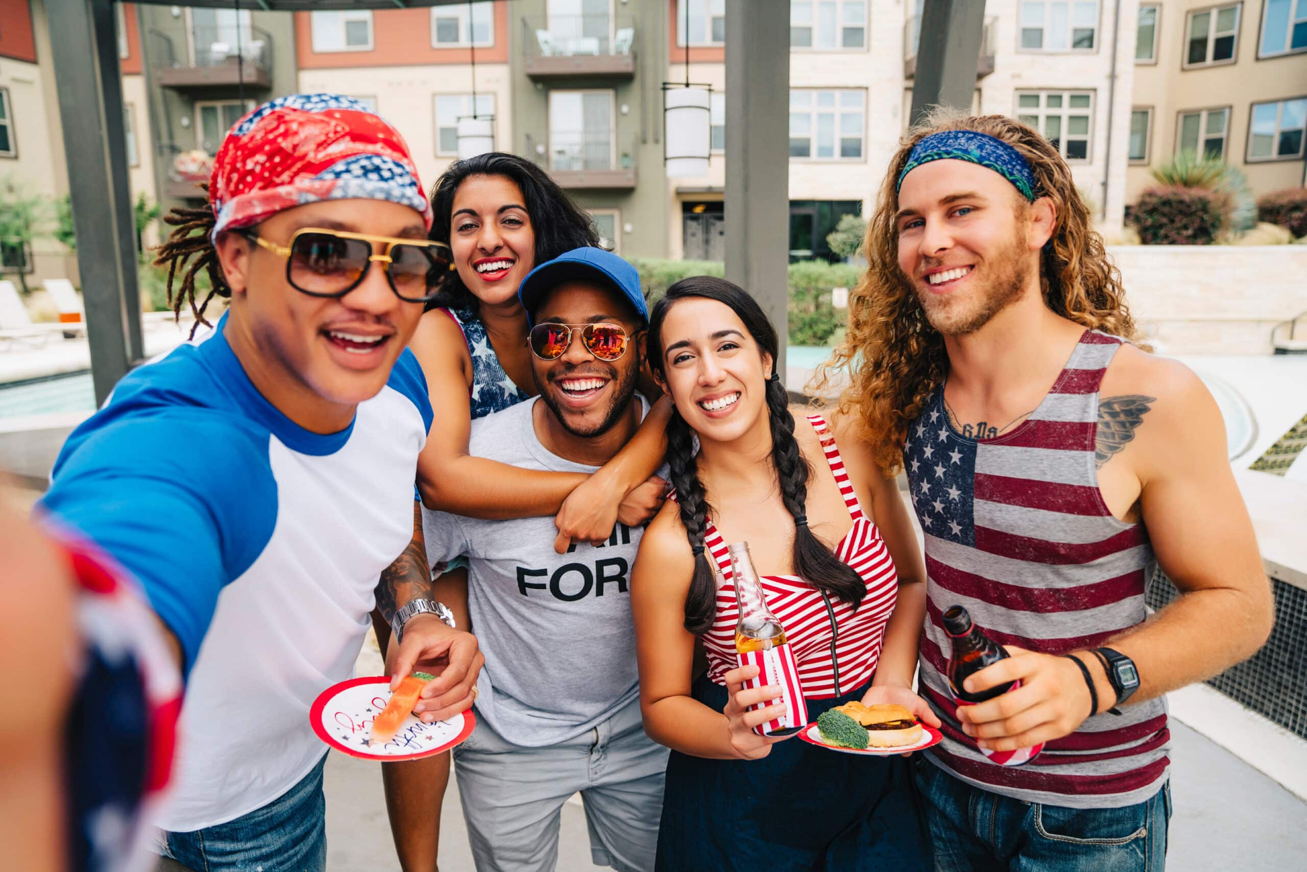 Group of five friends posing for a selfie at an outdoor pool, wearing red, white, and blue outfits and holding snacks/drinks in hand.