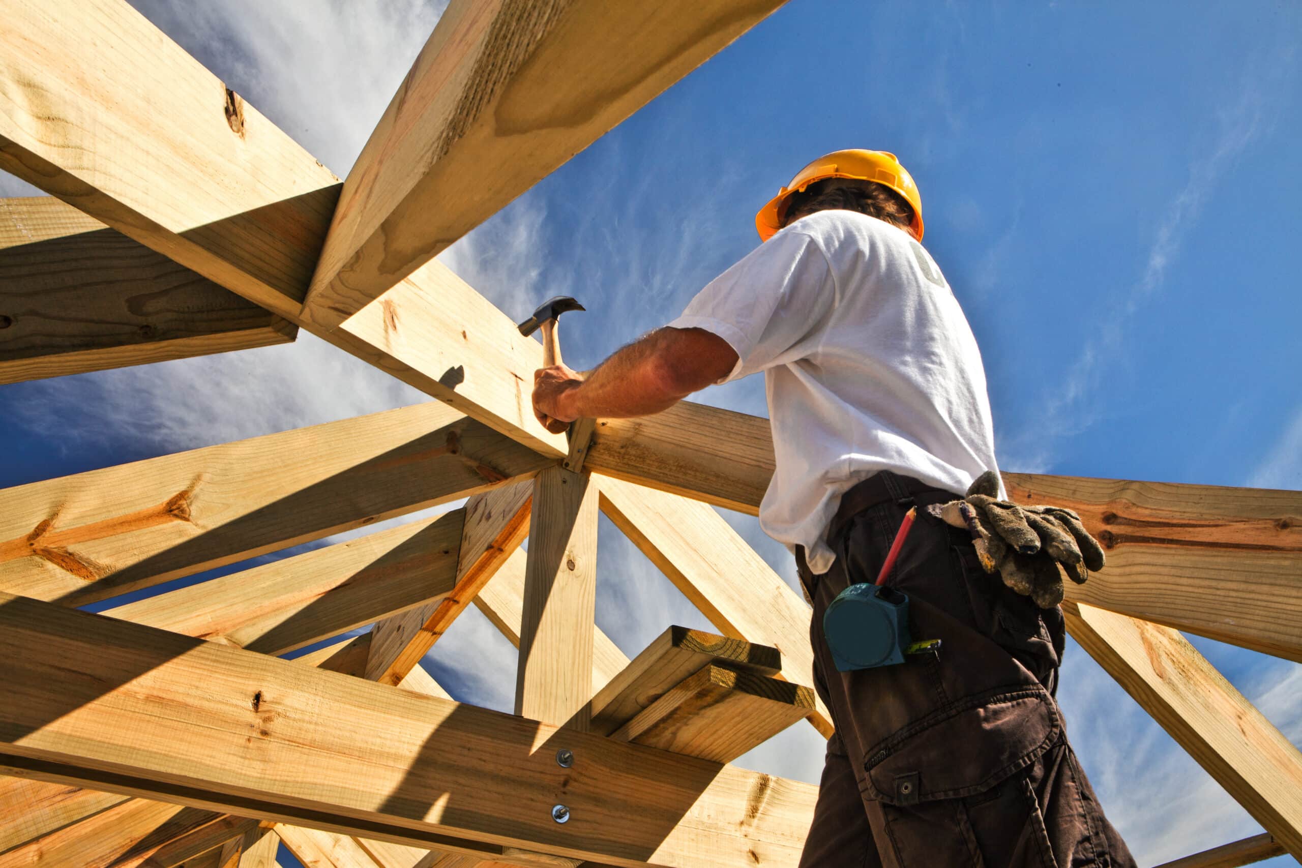 Construction worker wearing a yellow hard hat stands among wooden framing against a blue sky.