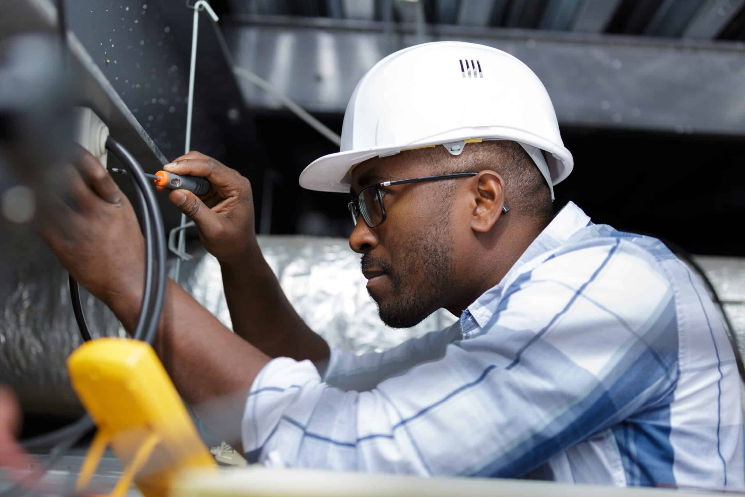 Worker wearing a white hard hat and safety glasses connects wires inside an industrial panel or equipment cabinet.